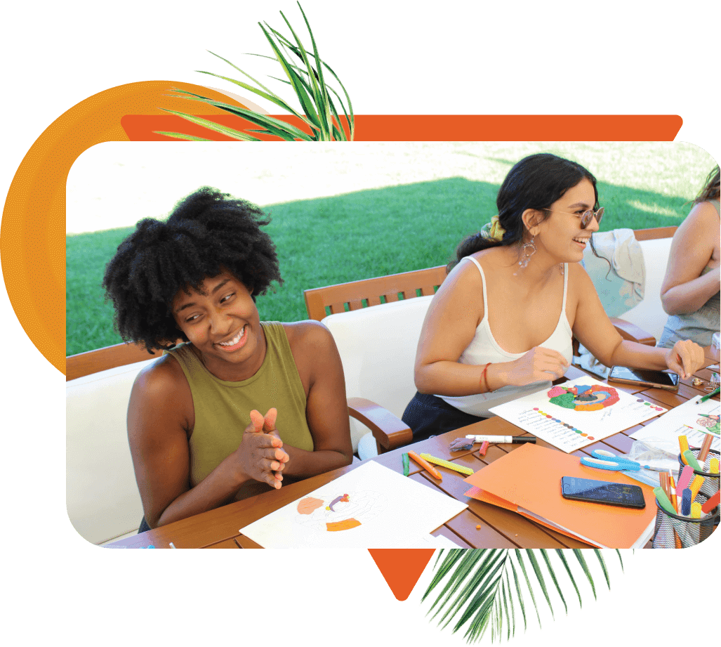 A young black woman and white woman seated at a table outdoors smiling during a workshop