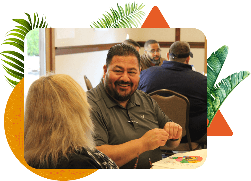 A middle-aged man with short dark hair and a beard smiling at a woman with blonde hair while sitting at a table during a workshop indoors