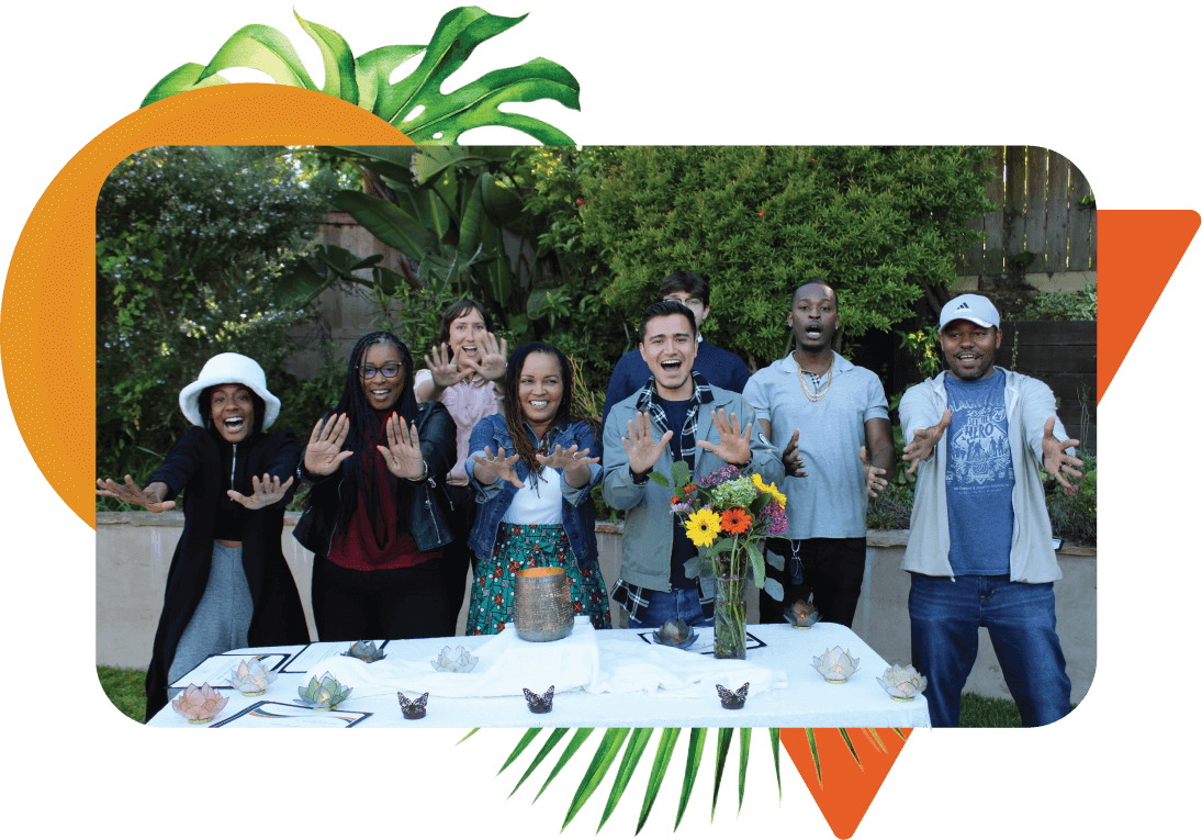 A diverse group of individuals standing together behind a table outdoors and smiling with their arms out toward the camera